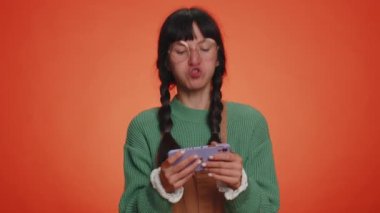 Endişeli, süveter giyen, cep telefonuyla video oyunu oynayan komik bağımlı kadın. Turuncu stüdyo arka planında izole edilmiş sürücü simülatörlü akıllı telefon uygulaması kullanan genç kız