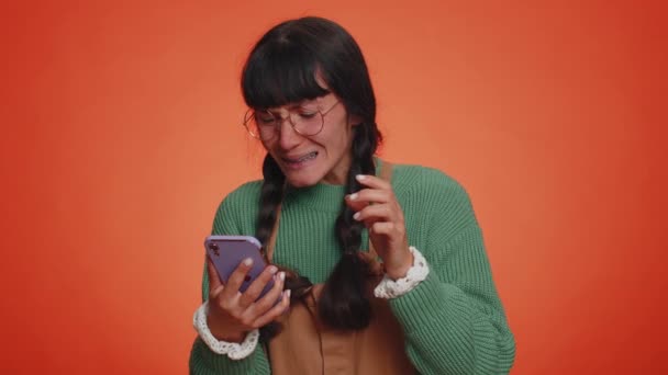 セーターのヒップスター女性は 携帯電話の入力ブラウジングを使用し 突然の宝くじの結果 失敗に驚いて失う 若いです女の子孤立した一人でオレンジスタジオの壁の背景 — ストック動画