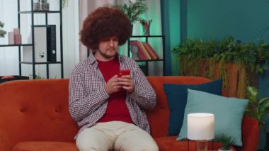Portrait of adult man sitting on sofa uses mobile phone smiles at modern home apartment. Young guy texting share messages content on smartphone social media applications online, watching relax movie