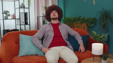 Repair work at neighbours concept. Irritated guy sits on couch cover ears with pillows annoyed by noisy neighbors suffer from headache wishes silence. Thin walls at home flat without sound insulation