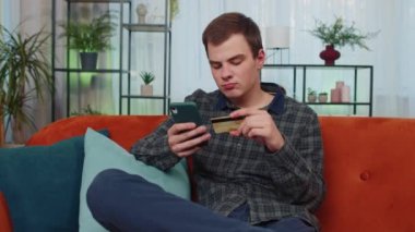 Kredi kartı ve akıllı telefon kullanan genç bir adam para transfer ederken, online alışveriş uygulaması satın alırken, ev içinde yemek siparişi verirken. Oturma odasında kanepede oturan genç adam.
