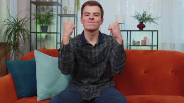 Happy caucasian man shouting, celebrating success, winning, goal achievement, good news, raising fists in gesture I did it, lottery luck at home apartment. Young guy indoors in living room on couch