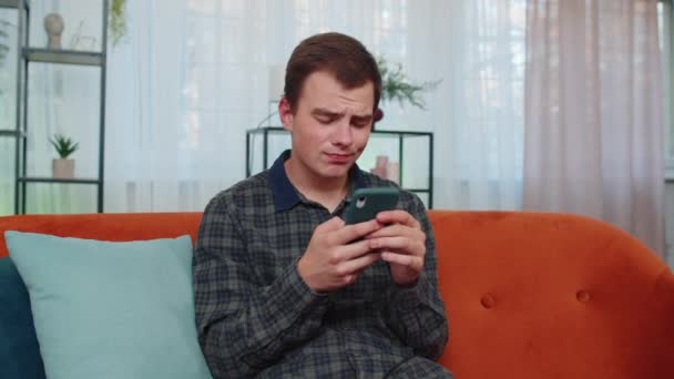 青少年男子的肖像用智能手机打字浏览 失去了突如其来的彩票结果 坏消息 财富损失 游戏失败 坐在家里客厅橙色沙发上的年轻人 — 图库视频影像