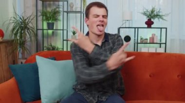 Coşkulu bir beyaz adam rock 'n roll jestini elleriyle yapıyor, havalı bir işaret veriyor, çılgın bir ifadeyle evet diye bağırıyor, dans ediyor, duygusal olarak başarıyla seviniyor. Evdeki genç adam.