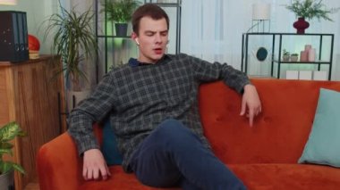 Happy relaxed overjoyed young guy in wireless earphones dancing on couch at home choosing listening favorite energetic disco rock n roll music in smartphone entertaining. People weekend activities