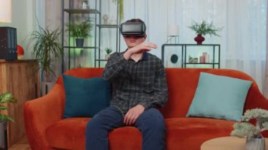 Genç adam evde VR kulaklık kullanıyor, 360 boyutlu video konseptinin tadını çıkarıyor, eğleniyor, olumlu ruh halinde, ellerini havada hareket ettiriyor, oturma odasında kanepede oturuyor. Sanal gerçeklik, modern aygıt