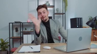 Businessman in suit working on laptop computer smiling friendly at camera and waving hands gesturing hello, hi, greeting or goodbye, welcoming with hospitable expression at office workplace desk
