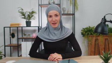 Başörtülü Müslüman iş kadını dizüstü bilgisayarda çalışıyor kameraya gülümsüyor ve selamlıyor, selamlıyor, selamlıyor, iş yerindeki çalışma masasında misafirperver bir ifadeyle karşılıyor.