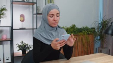 Modern ofis çalışma alanında geleneksel türban takan genç Müslüman iş kadını akıllı telefon sosyal medya uygulamalarındaki içeriği mesajlaşıyor, rahat film izliyor