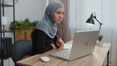 Kafiyeli çalışan Müslüman bir iş kadını dizüstü bilgisayarında not defteriyle web kamerasıyla konferans görüntülü konuşma yapıyor. Çevrimiçi öğretmen uzaktan sohbette e-öğreniyor. Çevrimiçi uzaktan eğitim