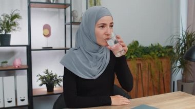 Susamış genç Müslüman kadın elinde bir bardak doğal su ile susuzluğu gidermek için bir yudum su içiriyor. Evde çalışan, iyi yaşam alışkanlıkları olan sağlıklı zayıflama konsepti olan bir kız.
