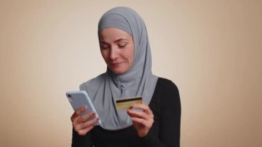 Para transferi, online alışveriş, ödeme yaparken kredi kartı ve akıllı telefon kullanan tesettürlü müslüman bir kadın. Finans ve internet. Bej arka planda izole edilmiş genç bir kız