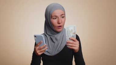 Tesettürlü mutlu müslüman kadın neşeli bir şekilde kazanıyor, nakit para alıyor, piyango talihi kazanıyor. Genç milenyum kızı bej renkli stüdyo geçmişinde izole edilmiş.