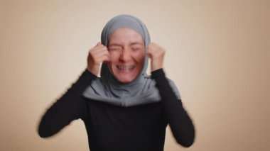 Mutlu, tesettürlü Müslüman kadın bağırıyor, el kol hareketi yapıyor başarıyı kutluyorum, kazanıyorum, doğum günü, piyango hedefine ulaşıyorum. Bej renkli stüdyo arka planında yetişkin bir Arap kız.