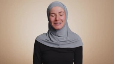 Başörtülü Müslüman iş kadını kameraya dostça gülümsüyor, onaylayarak başını sallıyor, merhaba işareti yapıyor, selamlıyor, misafirperver bir ifadeyle karşılıyor. Bej renkli stüdyo arka planında izole edilmiş Arap kız