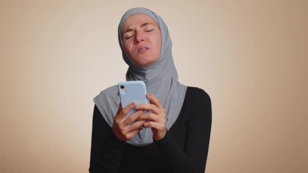 ヒジャーブのイスラム教徒の女性は 携帯電話の携帯電話の入力のブラウジングを使用すると 突然の宝くじの結果 失敗に驚いて失う 若いですアラビア語女の子絶縁だけでベージュスタジオ壁の背景 — ストック動画