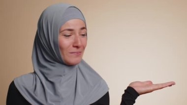Başparmak gösteren ve boş yerleri gösteren tesettürlü Müslüman bir kadın. Ticari metinler için reklam alanı, ürün tanıtımı için kopya alanı. Genç yetişkin bir kız. Bej duvar arka planında çekilen kapalı stüdyo.