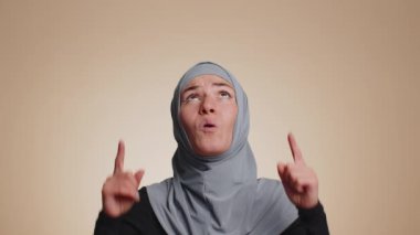 Başparmak gösteren ve boş yerleri gösteren tesettürlü Müslüman bir kadın. Ticari metinler için reklam alanı, ürün tanıtımı için kopya alanı. Genç yetişkin bir kız. Bej duvar arka planında çekilen kapalı stüdyo.