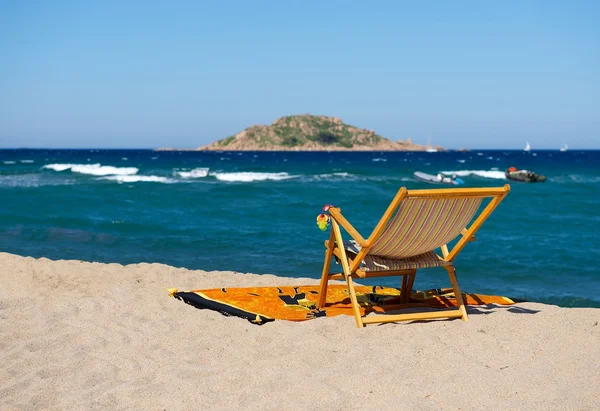 Strandkorb am gelben Sandstrand mit blauem Himmel, Strandkorb am Tag am Strand mit blauem Himmelshintergrund und verschwommenen Booten, kleine Insel weit weg, Sommerurlaub in Sardinen, ein Stuhl vor blauem Himmel — Stockfoto