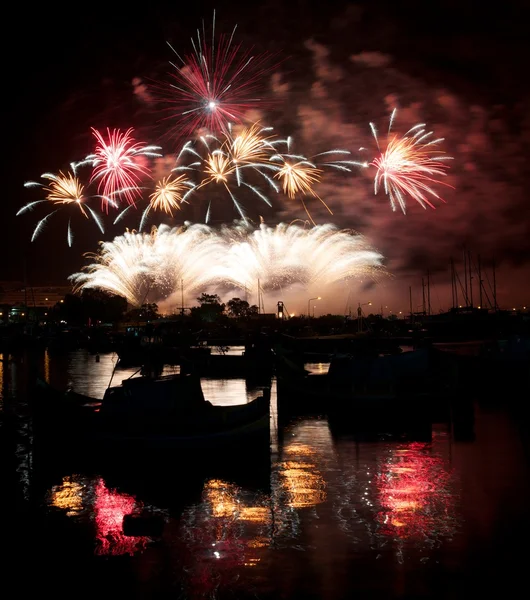 Fyrverkerier, explosion, fantastiska fyrverkerier med fin reflektion på ett vatten i malta, mörk himmel bakgrund och huset ljus i den avlägsna, malta fireworks festival, 4 juli självständighetsdagen — Stockfoto
