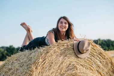 Gün batımında samanlıkta saman yığınında saman şapkalı siyah tulumlu güzel esmer kadın. Yüksek kalite fotoğraf