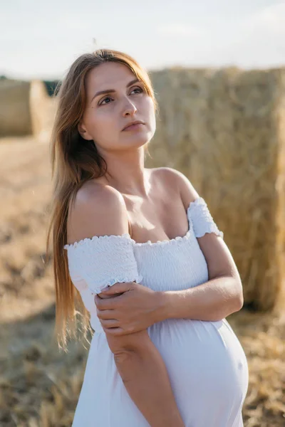 Gün batımında samanlığın yanındaki beyaz elbiseli güzel hamile esmer kadın. Yüksek kalite fotoğraf