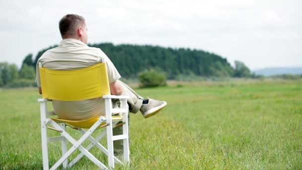 El hombre se sienta en una silla de picnic blanca y observa la naturaleza a su alrededor — Vídeos de Stock