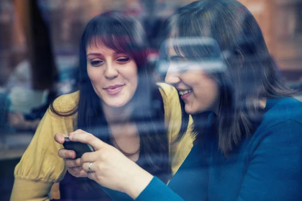 Dwie dziewczyny, uśmiechając się i za pomocą smartfona w kawiarni — Zdjęcie stockowe
