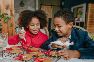 Kız ve erkek kardeş evde masada oturmuş zencefilli ekmek ve kurdele süslüyorlar. Noel zamanı aile eğlencesi konsepti. Bozuk el yapımı Mutlu Yıllar tatlısı, hediye ya da ağaç süslemesi fikri.