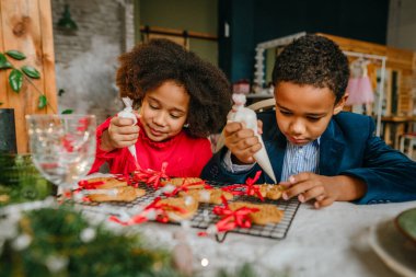 Kız ve erkek kardeş evde masada oturmuş zencefilli ekmek ve kurdele süslüyorlar. Noel zamanı aile eğlencesi konsepti. Bozuk el yapımı Mutlu Yıllar tatlısı, hediye ya da ağaç süslemesi fikri.