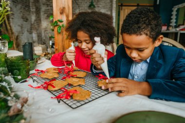 Kız ve erkek kardeş evde masada oturmuş zencefilli ekmek ve kurdele süslüyorlar. Noel zamanı aile eğlencesi konsepti. Bozuk el yapımı Mutlu Yıllar tatlısı, hediye ya da ağaç süslemesi fikri.