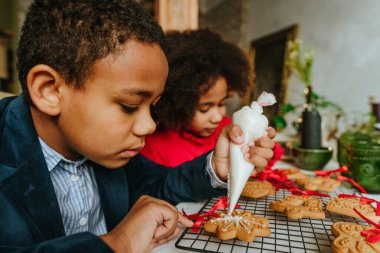 Kız ve erkek kardeş evde masada oturmuş zencefilli ekmek ve kurdele süslüyorlar. Noel zamanı aile eğlencesi konsepti. Bozuk el yapımı Mutlu Yıllar tatlısı, hediye ya da ağaç süslemesi fikri.