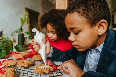 Kız ve erkek kardeş evde masada oturmuş zencefilli ekmek ve kurdele süslüyorlar. Noel zamanı aile eğlencesi konsepti. Bozuk el yapımı Mutlu Yıllar tatlısı, hediye ya da ağaç süslemesi fikri.
