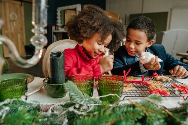 Kız ve erkek kardeş evde masada oturmuş zencefilli ekmek ve kurdele süslüyorlar. Noel zamanı aile eğlencesi konsepti. Bozuk el yapımı Mutlu Yıllar tatlısı, hediye ya da ağaç süslemesi fikri.