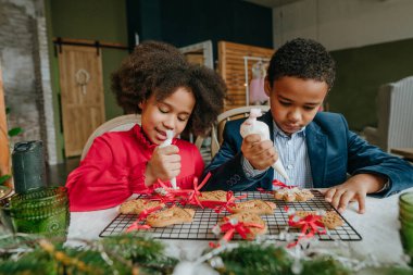 Kız ve erkek kardeş evde masada oturmuş zencefilli ekmek ve kurdele süslüyorlar. Noel zamanı aile eğlencesi konsepti. Bozuk el yapımı Mutlu Yıllar tatlısı, hediye ya da ağaç süslemesi fikri.