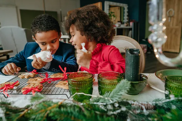 Kız ve erkek kardeş evde masada oturmuş zencefilli ekmek ve kurdele süslüyorlar. Noel zamanı aile eğlencesi konsepti. Bozuk el yapımı Mutlu Yıllar tatlısı, hediye ya da ağaç süslemesi fikri.