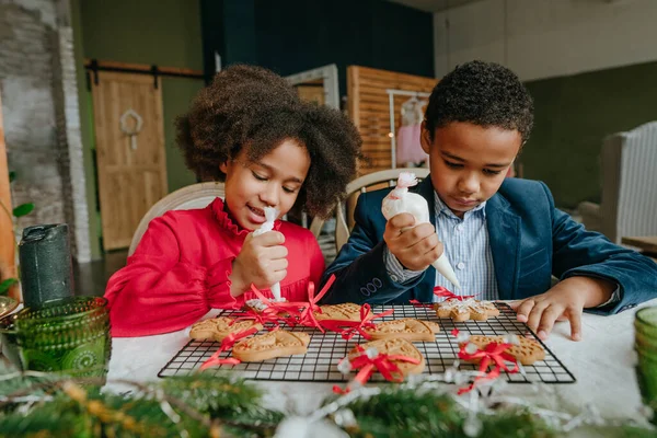 Kız ve erkek kardeş evde masada oturmuş zencefilli ekmek ve kurdele süslüyorlar. Noel zamanı aile eğlencesi konsepti. Bozuk el yapımı Mutlu Yıllar tatlısı, hediye ya da ağaç süslemesi fikri.