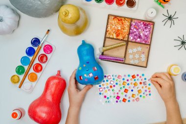 Boyalı balkabaklarını parlak çıkartmalarla boyayan çocuk. Çocuklar için cadılar bayramı sanat dersi fikri. Üst manzara, düz yatış. Mavi balkabağına odaklan.