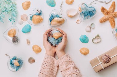 Beyaz arka planda deniz kabuğu ve yosun dolu süsler tutan kızların ellerini kapatın. Doğal malzemelerle Noel dekorasyonu fikri. Üst manzara, düz uzanma.