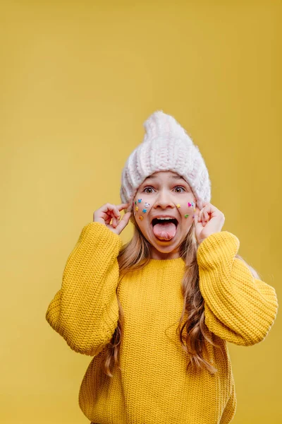 可爱的少女 脸上贴着贴纸 头戴粉色针织帽子 身穿黄色毛衣 在黄色背景上摆出舌头的样子 空白处有文字 — 图库照片