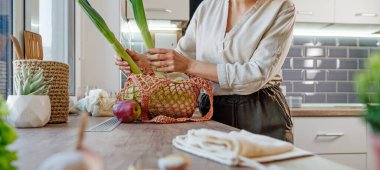 Mutfakta içinde meyve, sebze ve ekmek olan yeniden kullanılabilir ağ torbası tutan genç bir kadın. Sıfır atık ve sürdürülebilir ambalaj kavramı. Yatay panoramik pankart.
