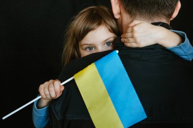 Karanlık arka planda 5 yaşında kızı olan bir baba. Ukrayna bayrağı taşıyan küçük bir kız babasına sarılıyor. Barış ve Ukrayna için dua et. Kızın yüzüne odaklan..