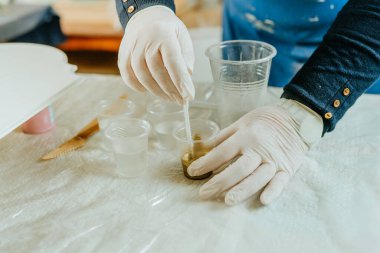 Resin sanat tekniğinde saat yapmak için malzemeleri hazırlayan sanatçı. Plastik eldiven giyen, altın tozlu pigmentle orta boyayı karıştıran bir kadın. Seçici odak.