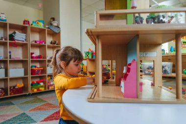 Anaokulunda ya da çocuk yuvasında oyuncak mobilyalarla ahşap evle oynayan küçük bir kız. Doğal malzeme oyuncakları ile erken gelişim. Montessori eğitim kavramı.