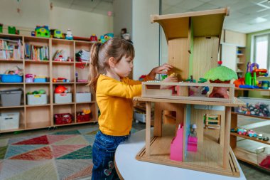 Anaokulunda ya da çocuk yuvasında oyuncak mobilyalarla ahşap evle oynayan küçük bir kız. Doğal malzeme oyuncakları ile erken gelişim. Montessori eğitim kavramı.