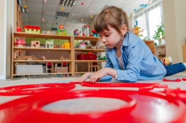 Anaokulunda ya da çocuk yuvasında yerde yatan kırmızı plastik oyuncaklarla oynayan küçük bir kız. Çocuk eğitimi konsepti.