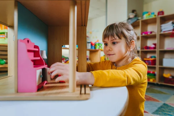 Little girl playing with wooden house with toy furniture at kindergarten or childcare centre. Early development with natural material toys. Montessori education concept.