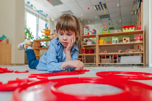Anaokulunda ya da çocuk yuvasında yerde yatan kırmızı plastik oyuncaklarla oynayan küçük bir kız. Çocuk eğitimi konsepti.