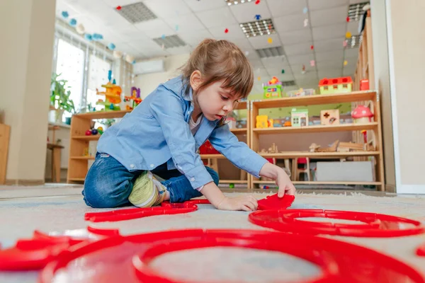 Anaokulunda ya da çocuk yuvasında yerde oturan kırmızı plastik oyuncaklarla oynayan küçük bir kız. Çocuk eğitimi konsepti.