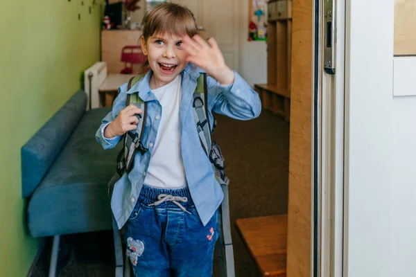 First day at school or kindergarten. Happy little girl with backpack standing near the door at school waving her hand. Back to school concept.